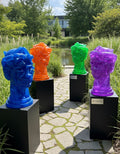 Four colorful sculptures of heads on stands in a garden setting with a pond and trees. Artwork by Mr. Clever Art.