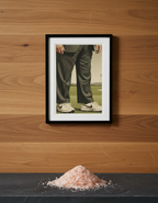 Framed artwork of a person's legs in pants and shoes on a black wall with pink salt in front.