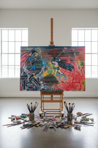 A large colorful canvas painting on an easel in a bright studio, showing a comic-style battle scene. Paintbrushes and paint containers are scattered on the floor around the easel.